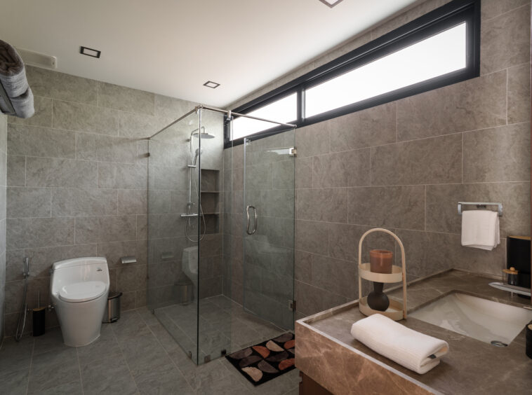 Modern bathroom with glass-walled shower, grey tile walls, toilet, and a bathtub; high horizontal window above shower.
