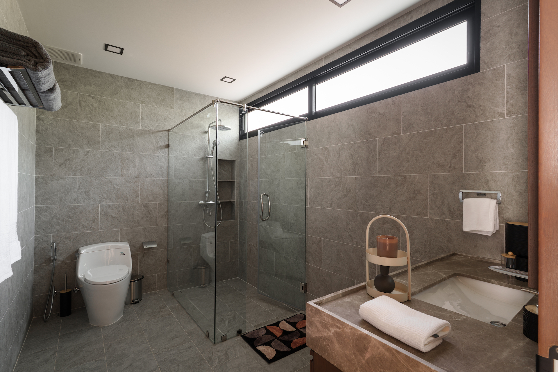 Modern bathroom with glass-walled shower, grey tile walls, toilet, and a bathtub; high horizontal window above shower.