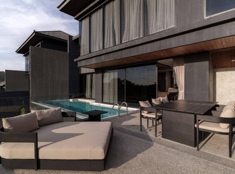 Modern outdoor lounge beside a rectangular pool on a glass-railed terrace with large windows and dark exterior.