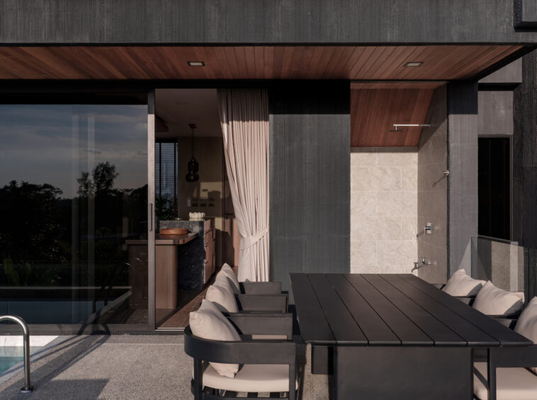 Modern outdoor dining area on a terrace with a dark wooden table and cushioned chairs, beside a glass-walled interior.