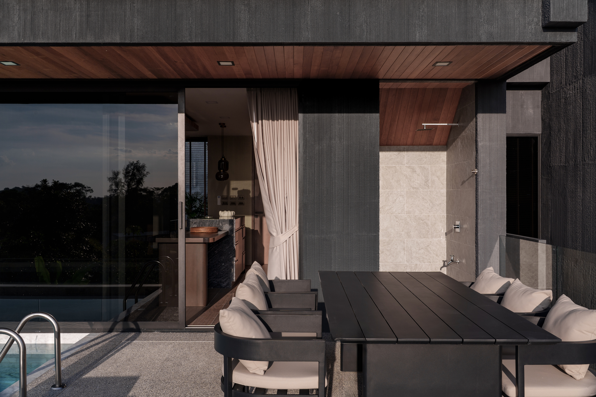 Modern outdoor dining area on a terrace with a dark wooden table and cushioned chairs, beside a glass-walled interior.