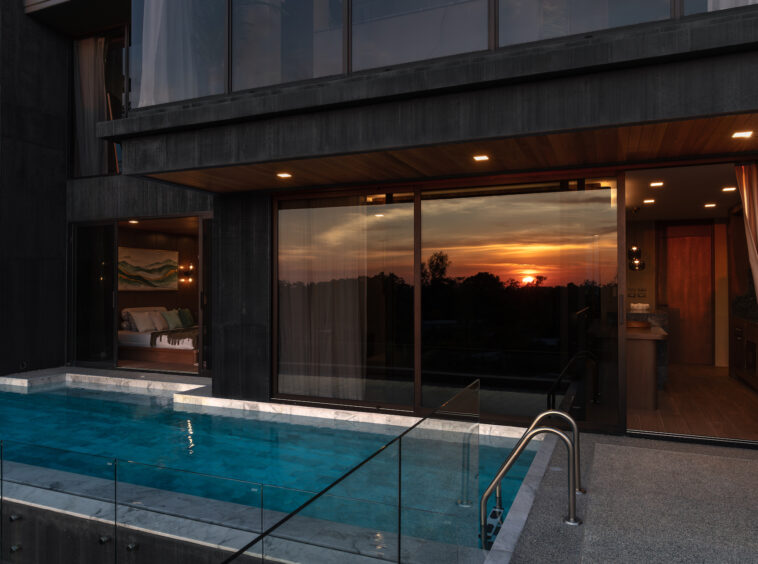 Modern dark-clad house with a glass-walled bedroom opening to a rectangular pool at sunset.
