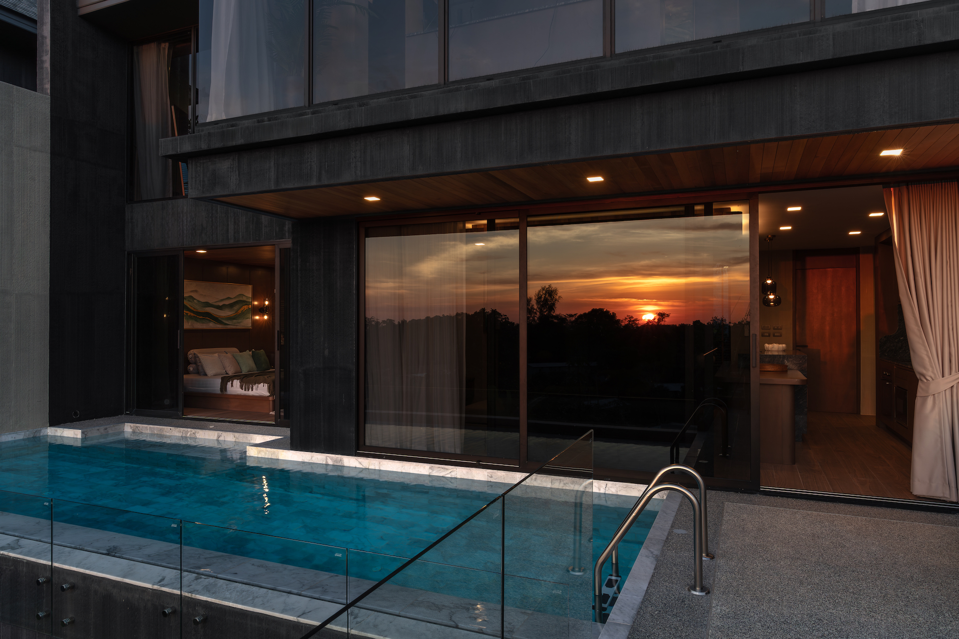 Modern dark-clad house with a glass-walled bedroom opening to a rectangular pool at sunset.