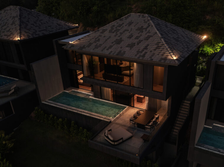 Aerial view of a modern hillside villa at night with warm-lit interiors and two rectangular pools.