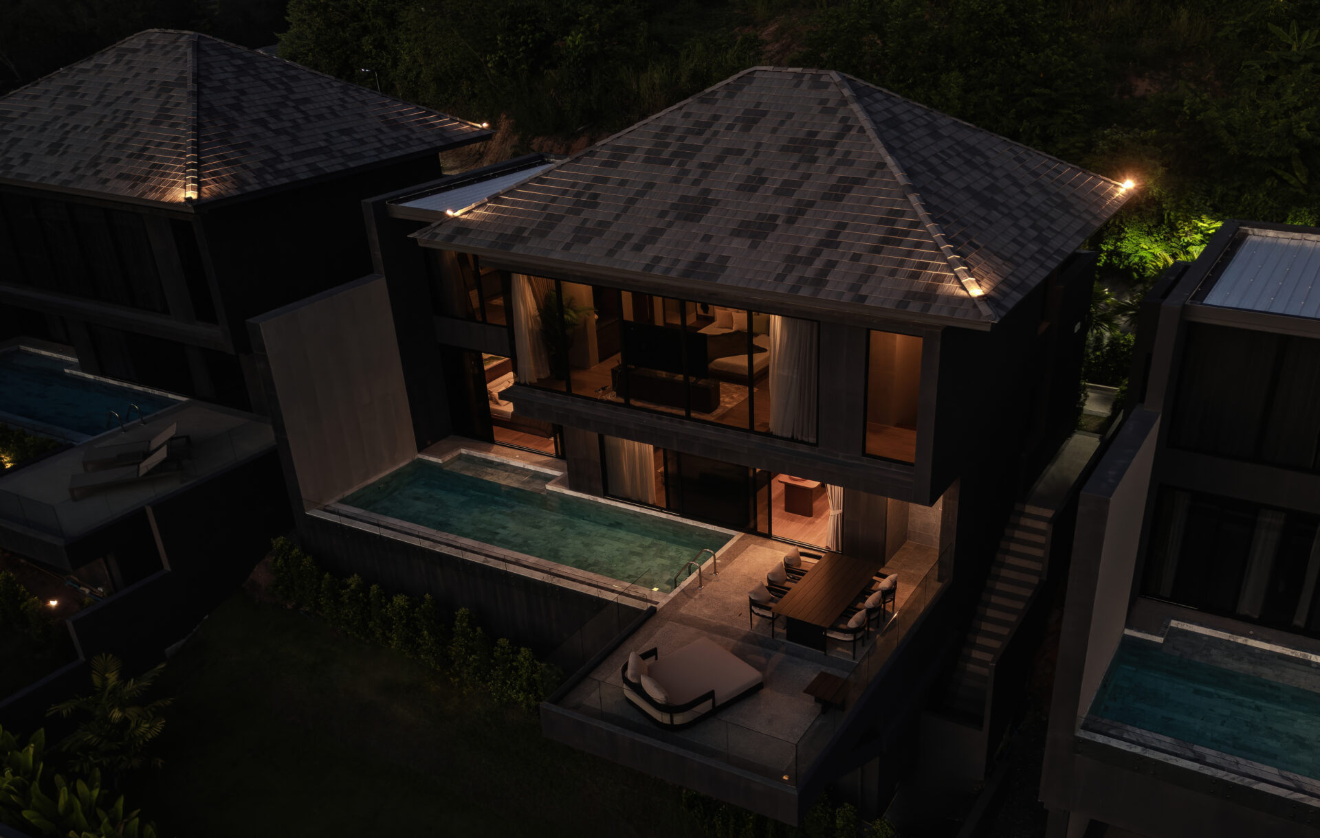 Aerial view of a modern hillside villa at night with warm-lit interiors and two rectangular pools.