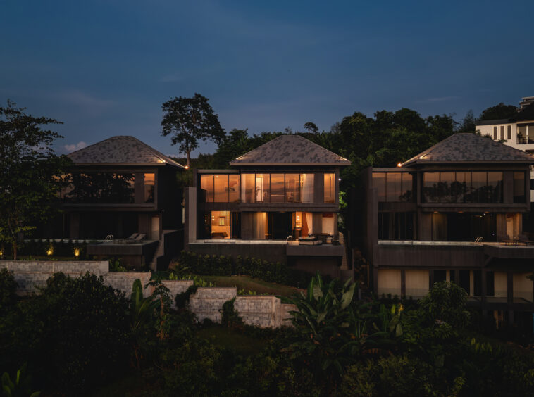 Row of modern glass-walled houses at dusk with warm interior lighting and tropical greenery in the foreground.