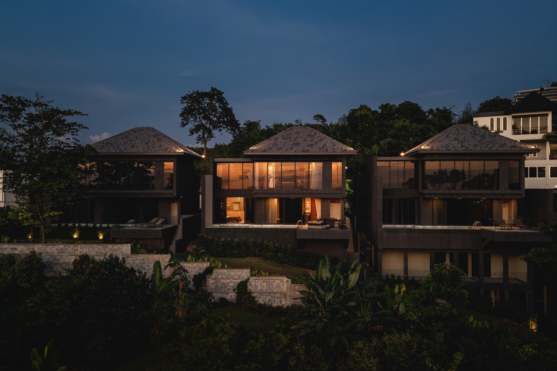 Row of modern glass-walled houses at dusk with warm interior lighting and tropical greenery in the foreground.