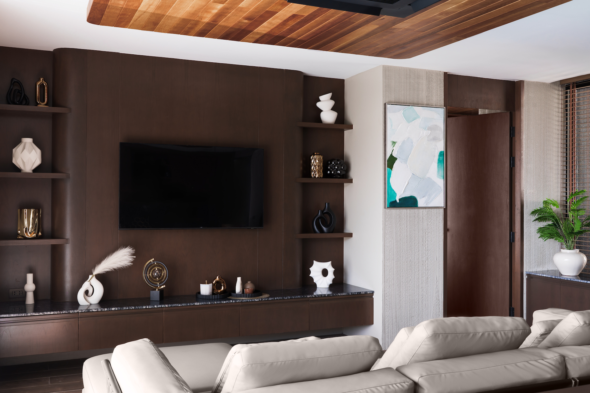 Modern living room with dark wood walls, wall-mounted TV, open shelves of vases and sculptures, and a beige sectional facing the screen.