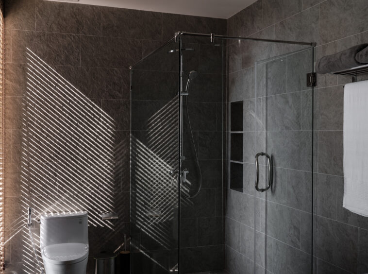 Modern bathroom with glass shower enclosure, dark tiled walls, and a white toilet; sunlight casts diagonal blinds shadows on the wall.