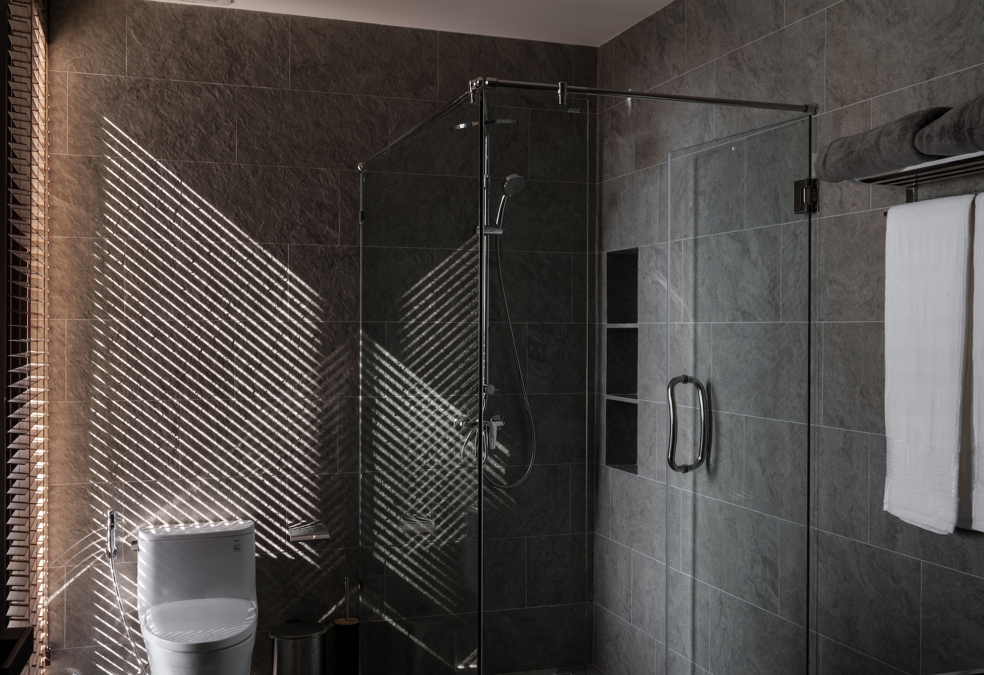 Modern bathroom with glass shower enclosure, dark tiled walls, and a white toilet; sunlight casts diagonal blinds shadows on the wall.