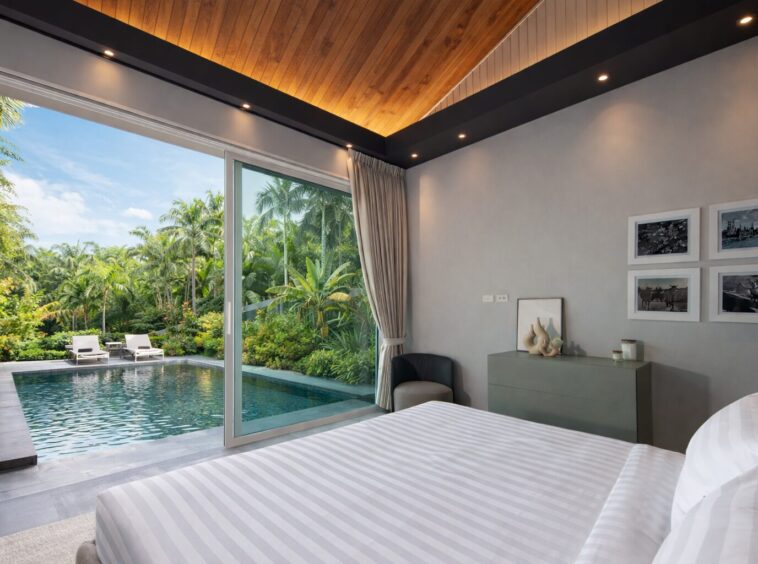 Bedroom with a large sliding door opening to a tropical pool and garden view; bed in foreground and modern dresser against gray wall.