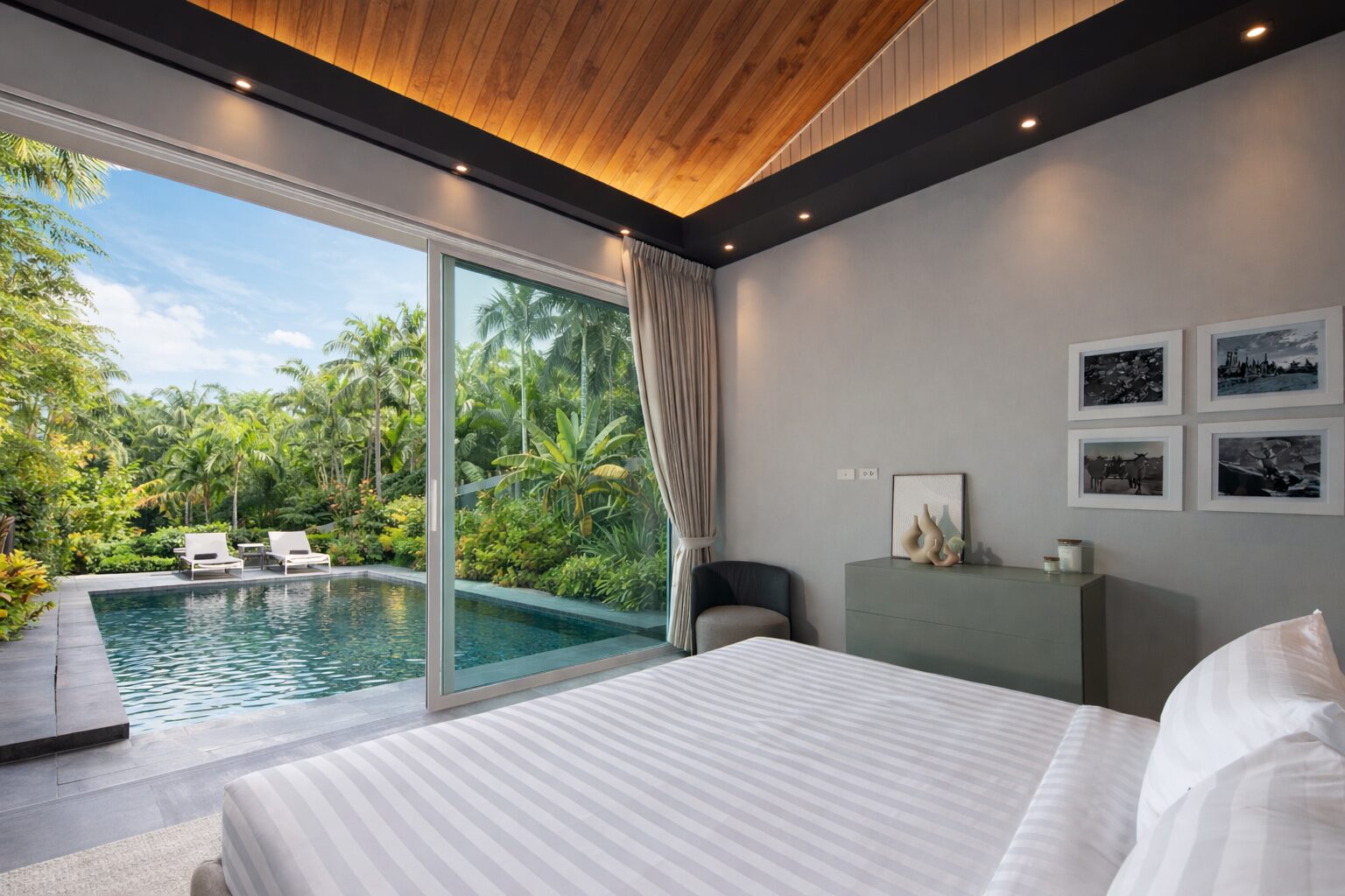 Bedroom with a large sliding door opening to a tropical pool and garden view; bed in foreground and modern dresser against gray wall.