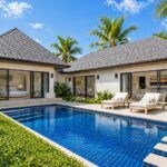 Luxury villa with a blue-tiled pool, two sun loungers, and palm trees in a tropical courtyard.