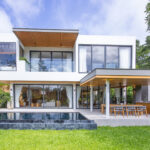 Modern two-story white house with glass walls, an outdoor dining area and a rectangular pool in the foreground.