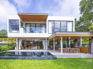 Modern two-story white house with glass walls, an outdoor dining area and a rectangular pool in the foreground.