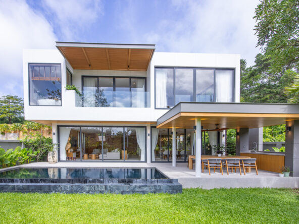 Modern two-story white house with glass walls, an outdoor dining area and a rectangular pool in the foreground.