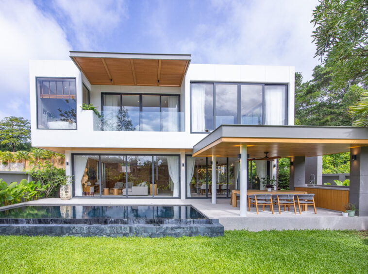 Modern two-story white house with glass walls, an outdoor dining area and a rectangular pool in the foreground.