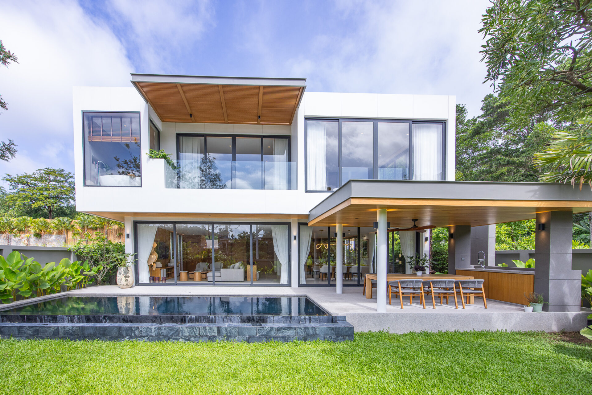 Modern two-story white house with glass walls, an outdoor dining area and a rectangular pool in the foreground.