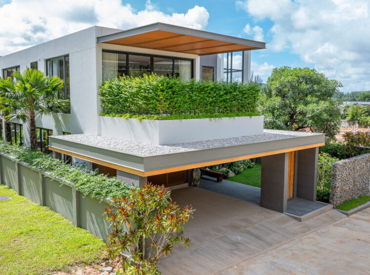 Modern white two-story house with large windows and a planted terrace, set among lush greenery and a concrete driveway under a blue sky.