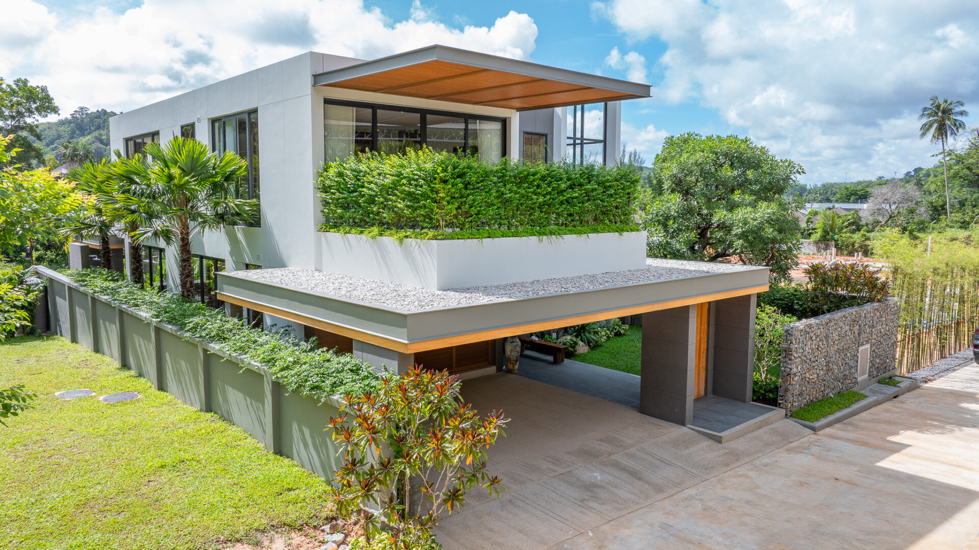 Modern white two-story house with large windows and a planted terrace, set among lush greenery and a concrete driveway under a blue sky.