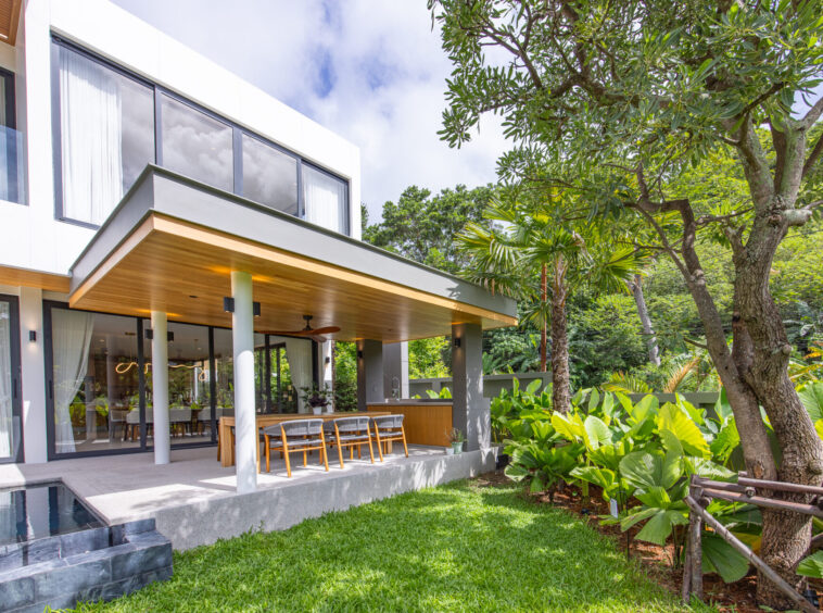 Contemporary home with glass walls, a covered patio dining area, and tropical landscaping including trees and shrubs.