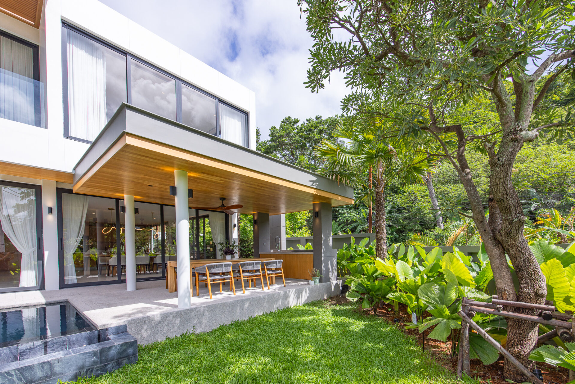Contemporary home with glass walls, a covered patio dining area, and tropical landscaping including trees and shrubs.