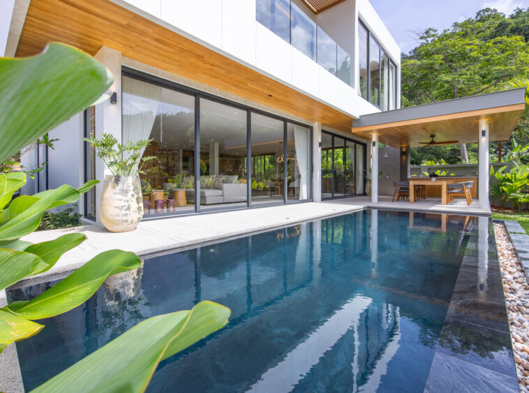 Modern two-story house with floor-to-ceiling glass doors, wooden overhang, and an in-ground pool on a sunny day.