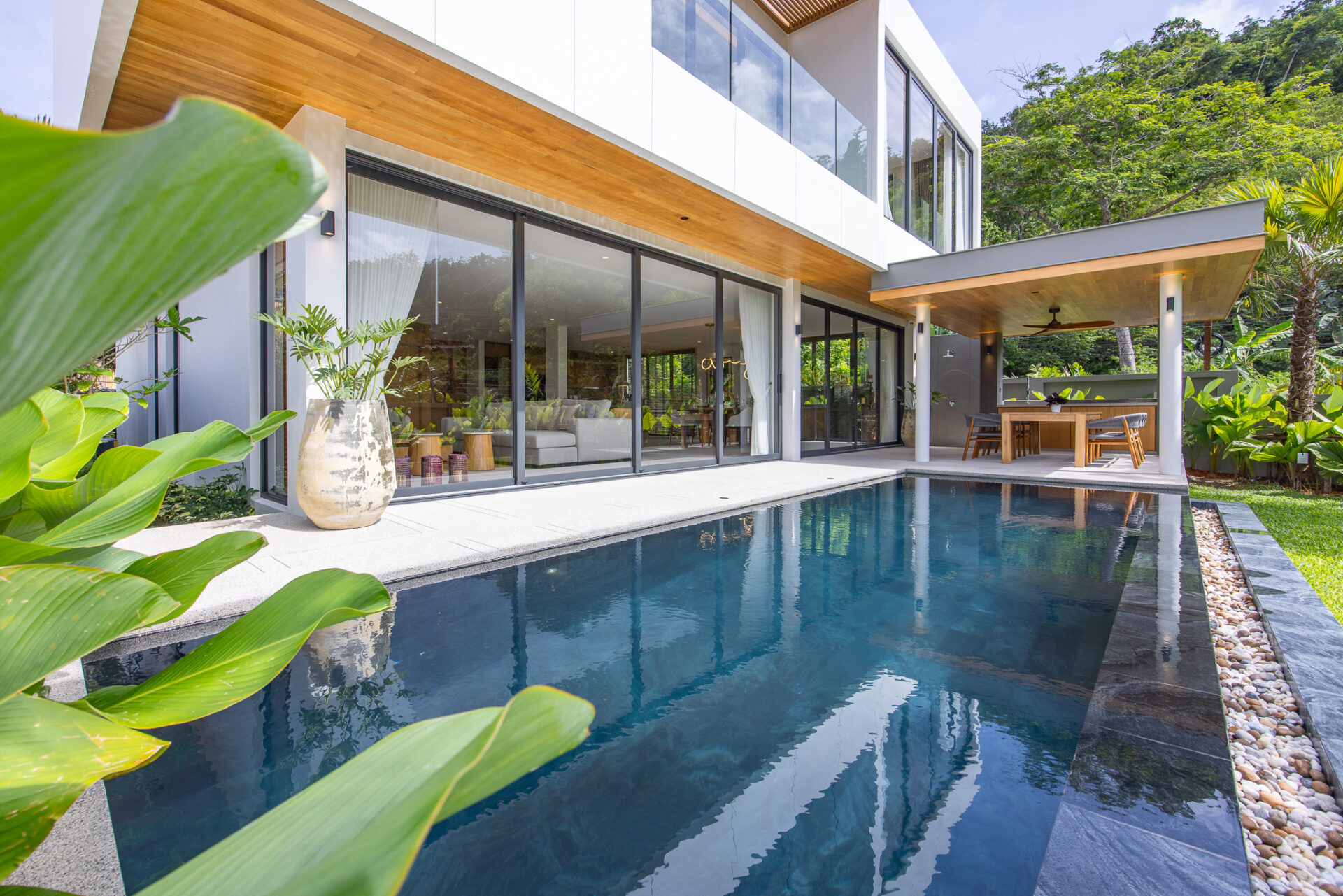 Modern two-story house with floor-to-ceiling glass doors, wooden overhang, and an in-ground pool on a sunny day.