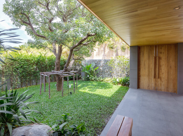 Modern covered patio with wooden double doors, gray tile floor, and a lush garden featuring a mature tree and greenery