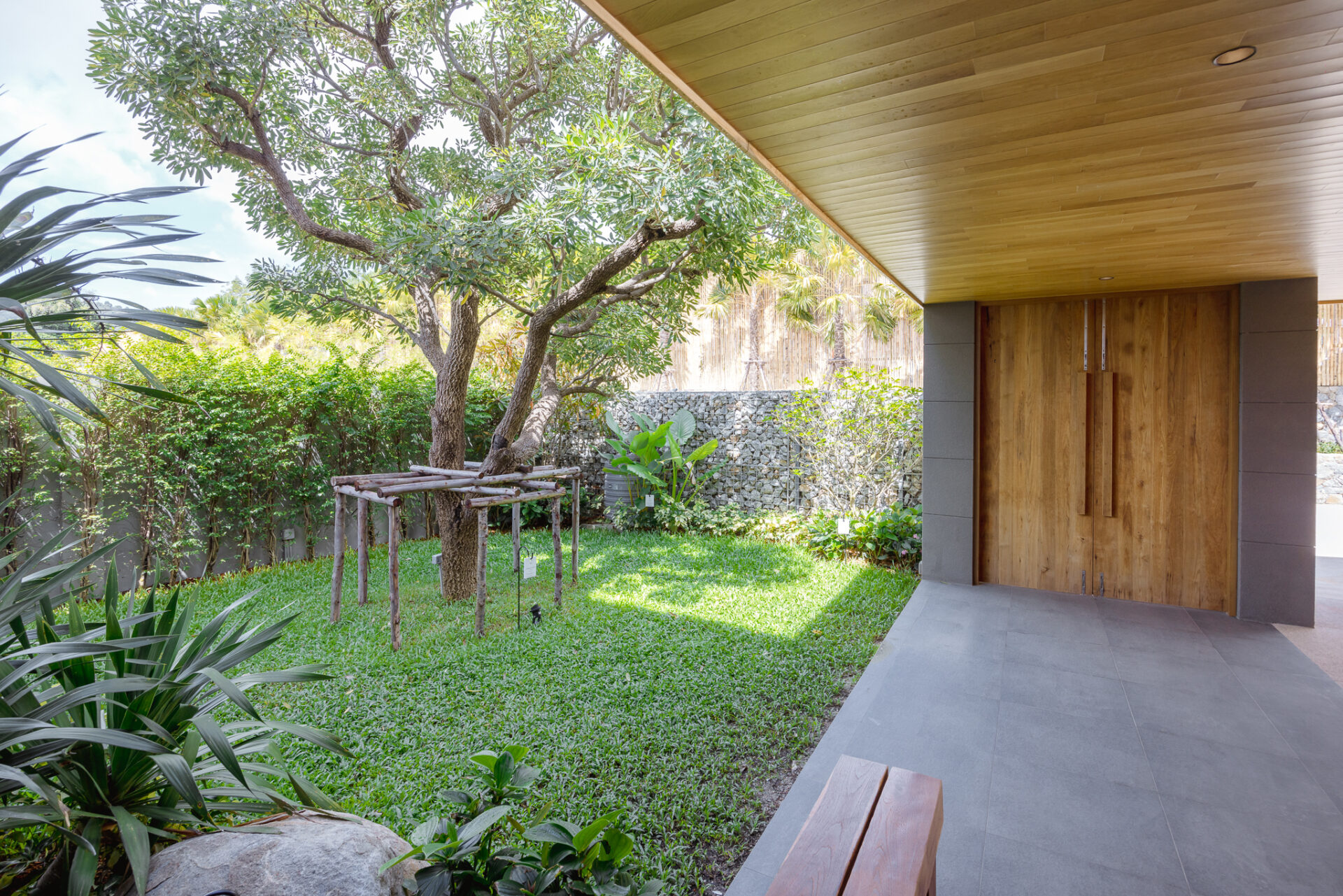 Modern covered patio with wooden double doors, gray tile floor, and a lush garden featuring a mature tree and greenery