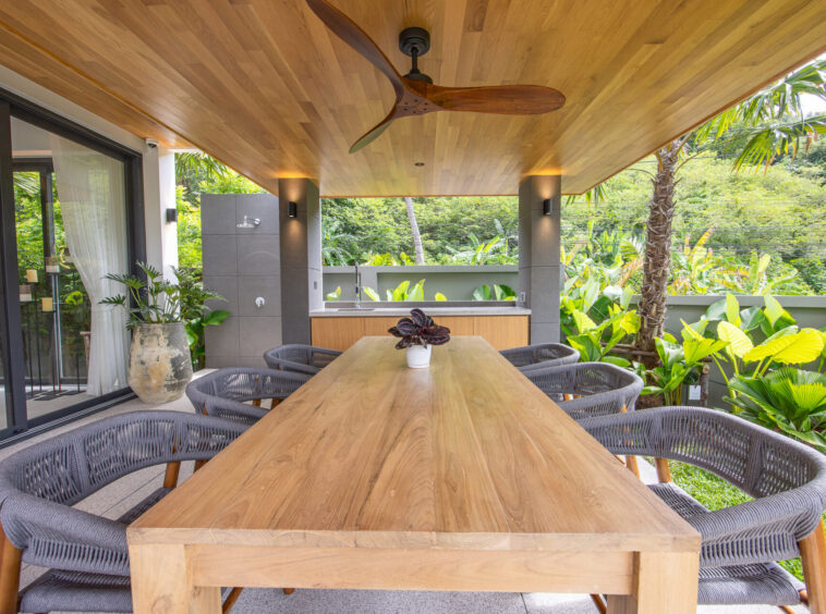 Outdoor covered patio with a long wooden dining table and wicker chairs, surrounded by tropical plants and a wood-paneled ceiling with a ceiling fan.