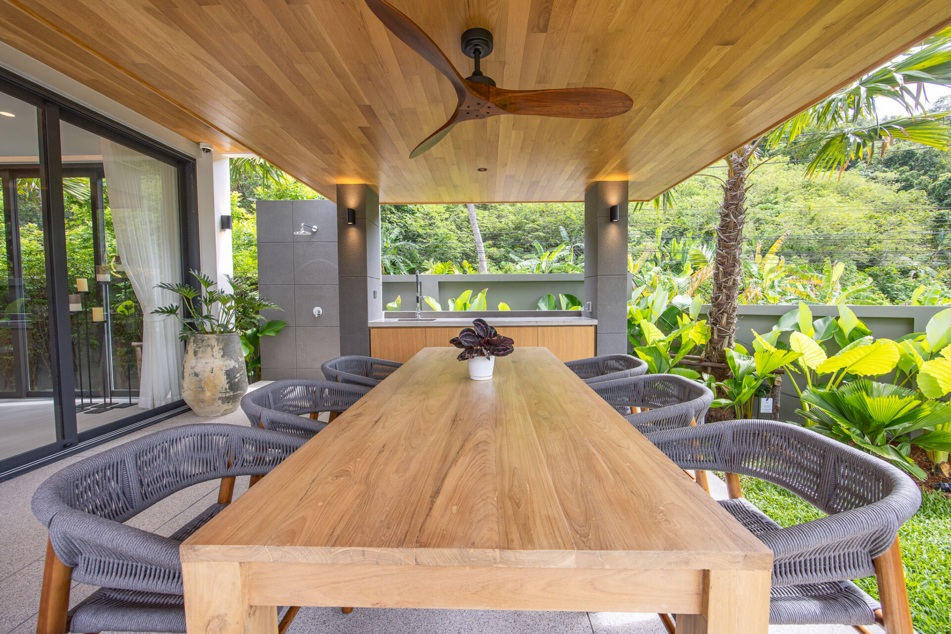 Outdoor covered patio with a long wooden dining table and wicker chairs, surrounded by tropical plants and a wood-paneled ceiling with a ceiling fan.