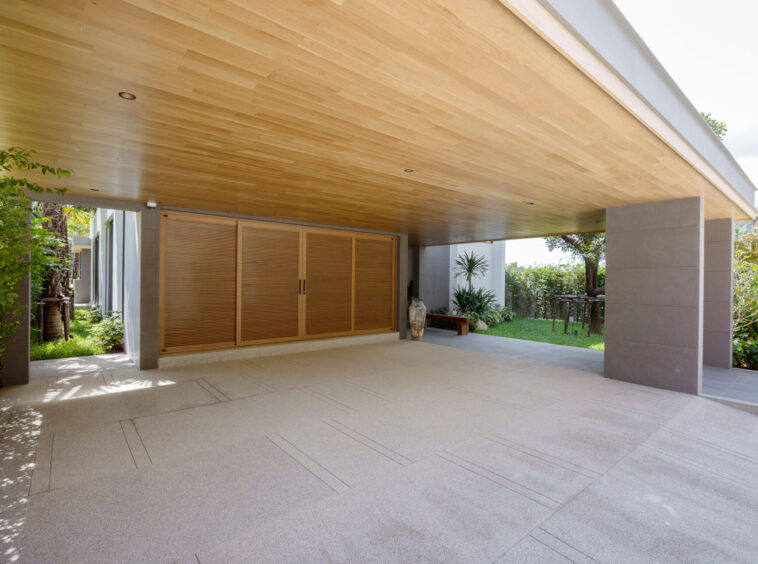 Covered outdoor patio with a warm wood-paneled ceiling, beige tiled floor, and large wooden shutters; plants and lawn beyond.