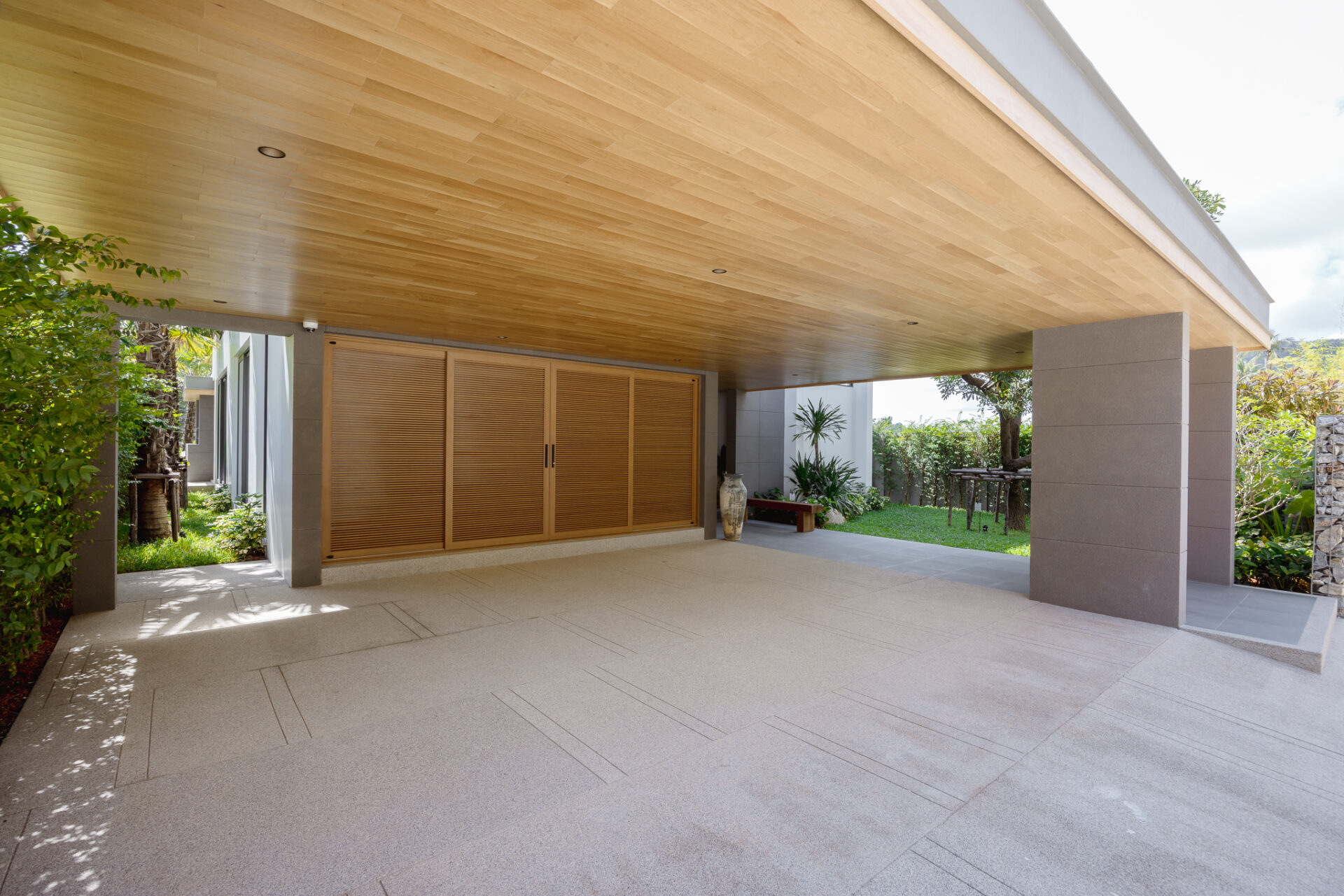 Covered outdoor patio with a warm wood-paneled ceiling, beige tiled floor, and large wooden shutters; plants and lawn beyond.