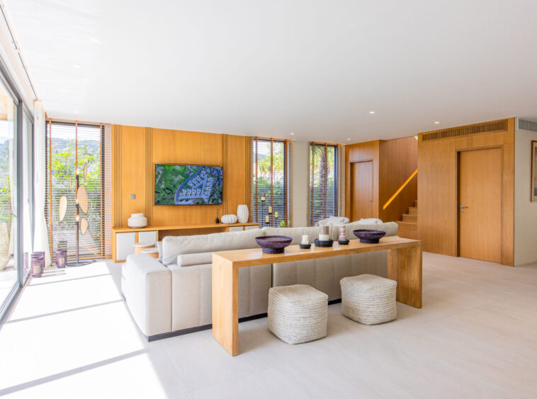 Bright open-concept living room with a beige sectional, wooden walls, and large glass doors to the outdoors. Whether informative or decorative, it conveys a spacious, sunlit living area.