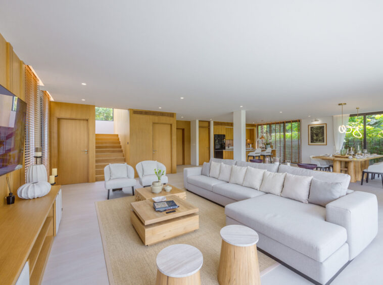 Open-concept living room with a light gray sectional, wood-paneled walls, and a dining area by large windows.