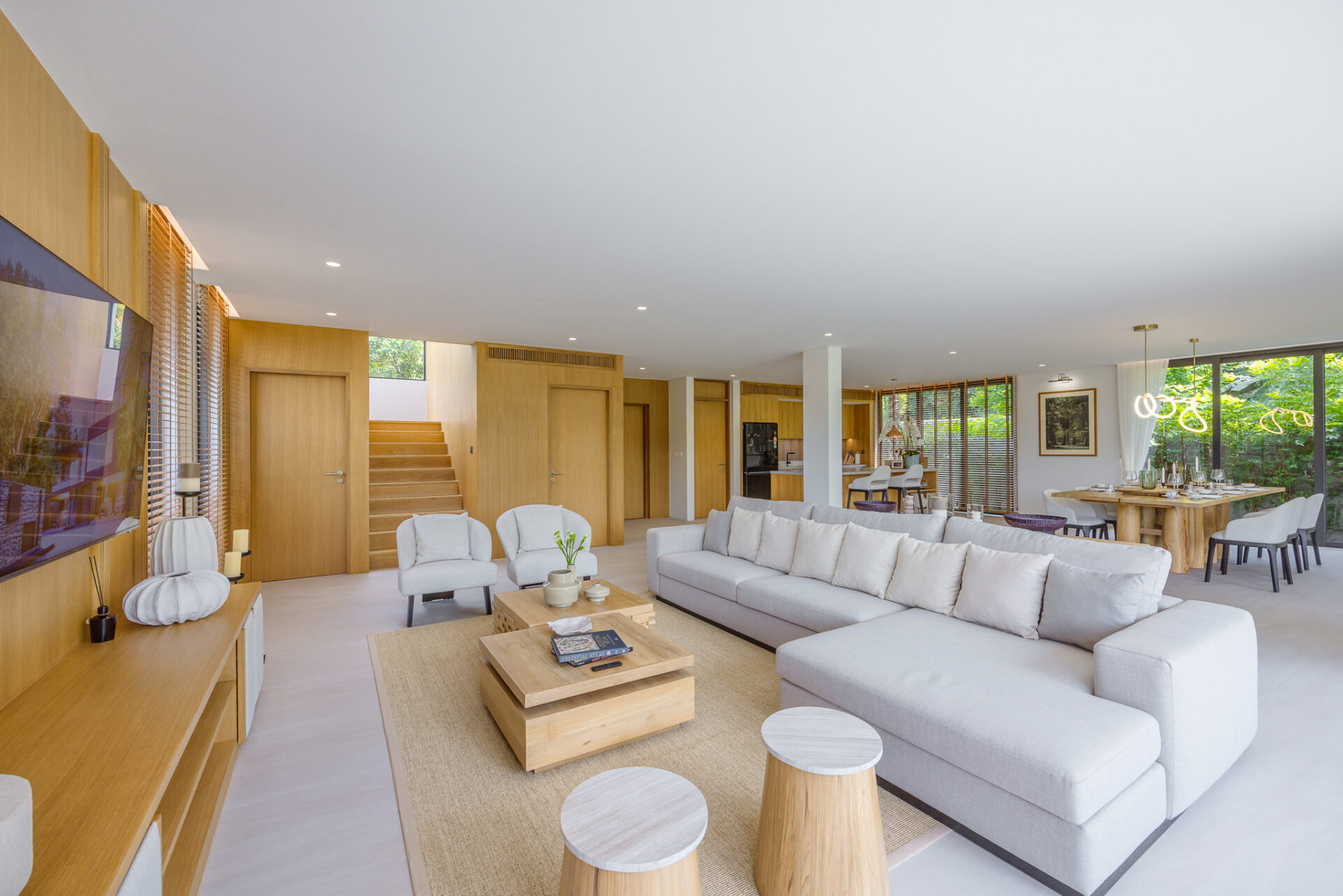 Open-concept living room with a light gray sectional, wood-paneled walls, and a dining area by large windows.