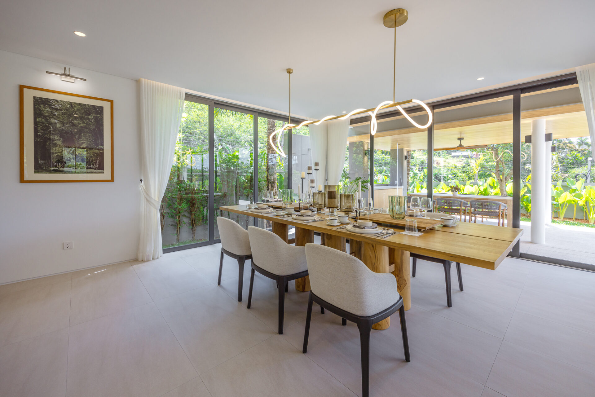 Bright dining room with a long wooden table set for a meal, white upholstered chairs, and a sculptural wavy pendant light above.