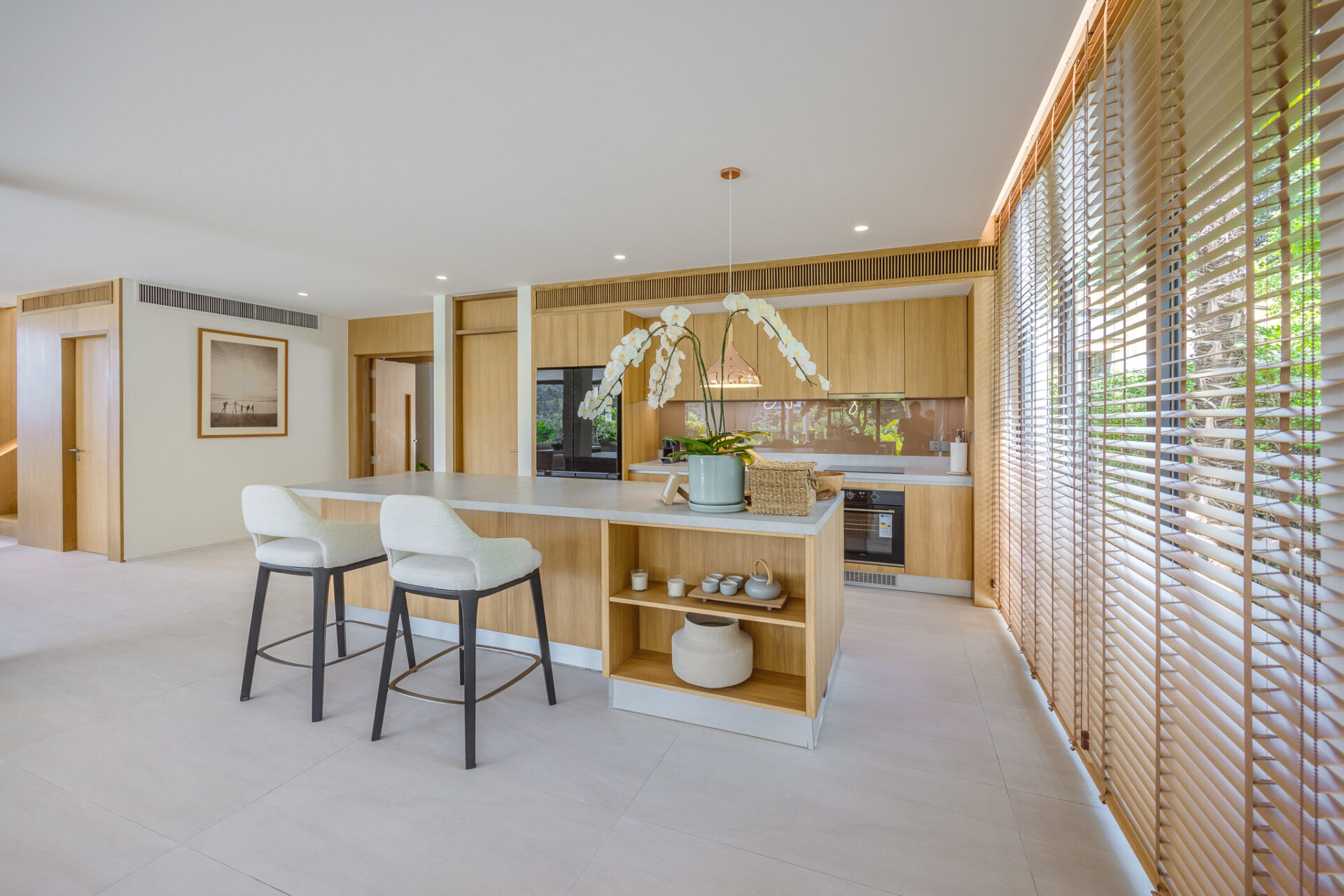 Modern kitchen with a central island, three cream stools, and large wooden‑louvered windows','Open-plan kitchen featuring oak cabinets, built-in oven, and an orchid on the island' ,
