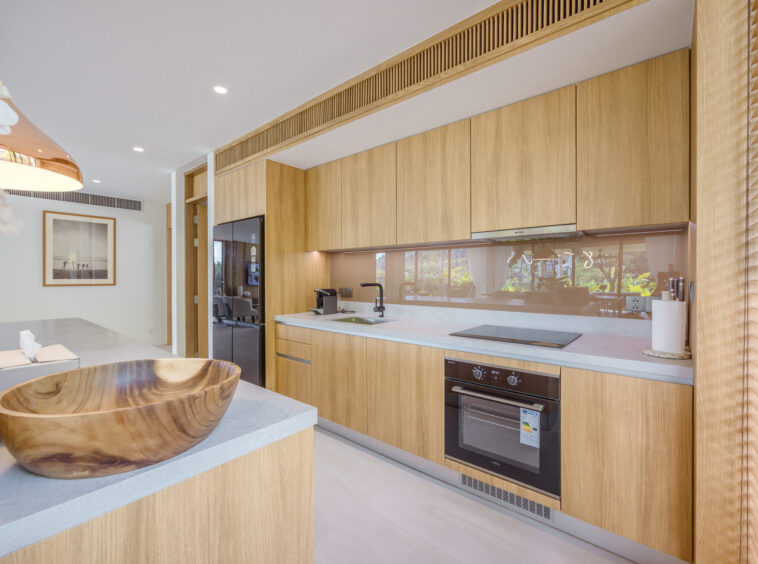 Modern kitchen with light wood cabinets, white countertops, and a built-in oven along the wall; large window blinds on the right.