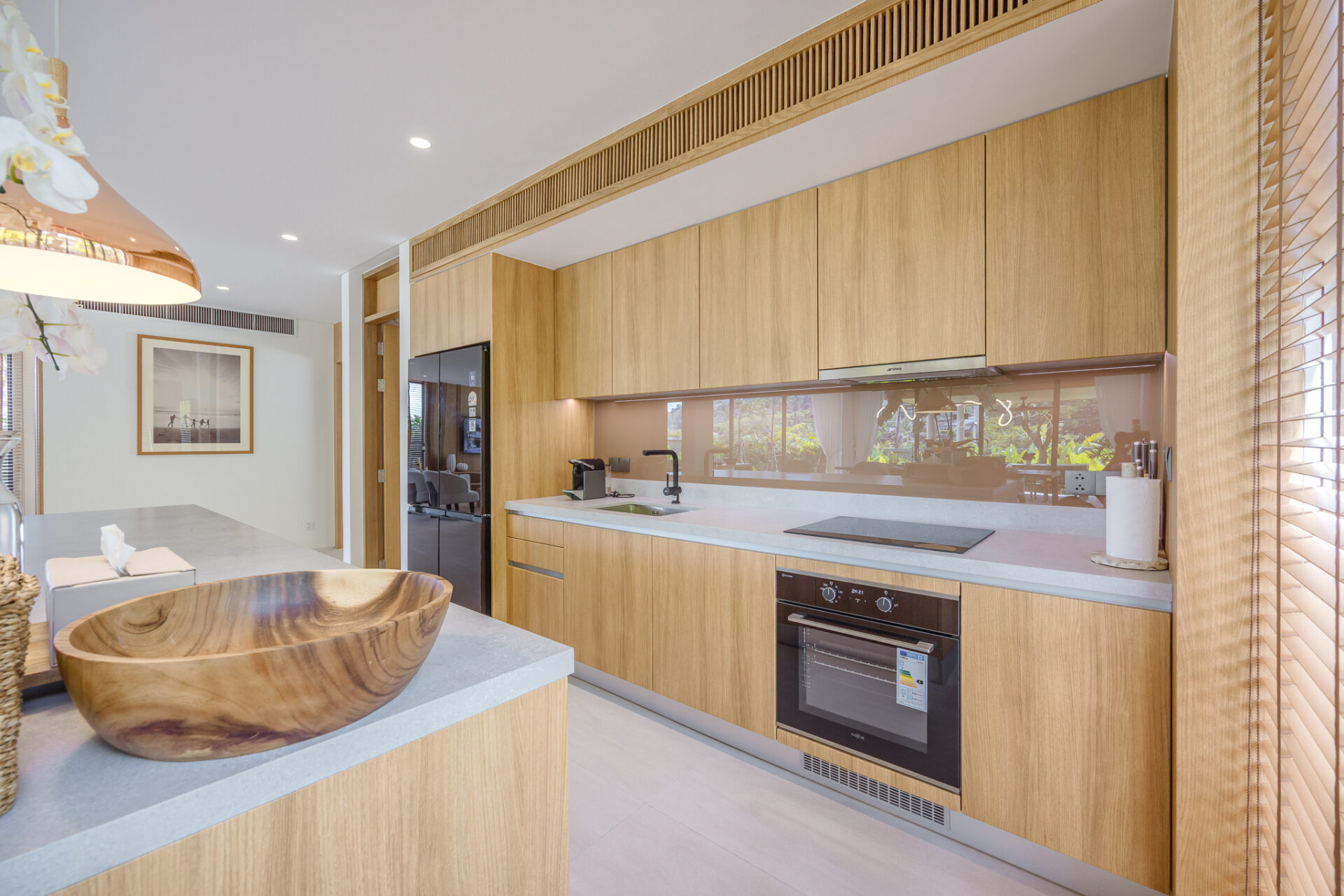 Modern kitchen with light wood cabinets, white countertops, and a built-in oven along the wall; large window blinds on the right.