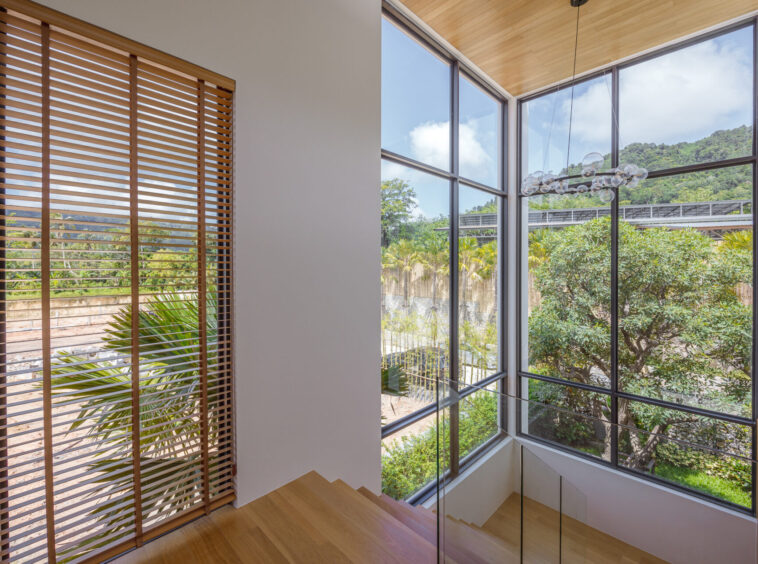 Staircase by large corner windows with wooden blinds; natural light and greenery outside the patio area nearby a glass railing.