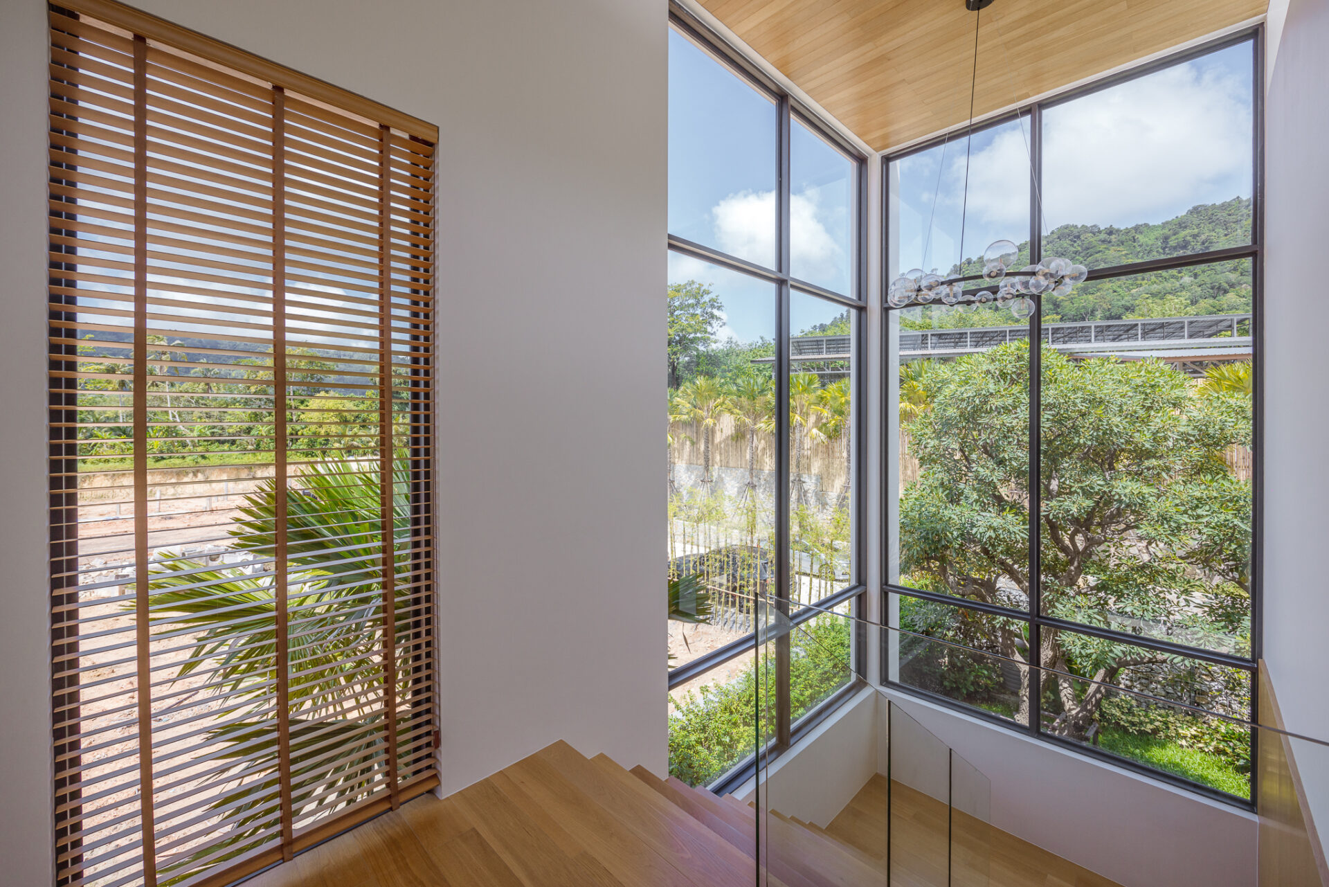 Staircase by large corner windows with wooden blinds; natural light and greenery outside the patio area nearby a glass railing.