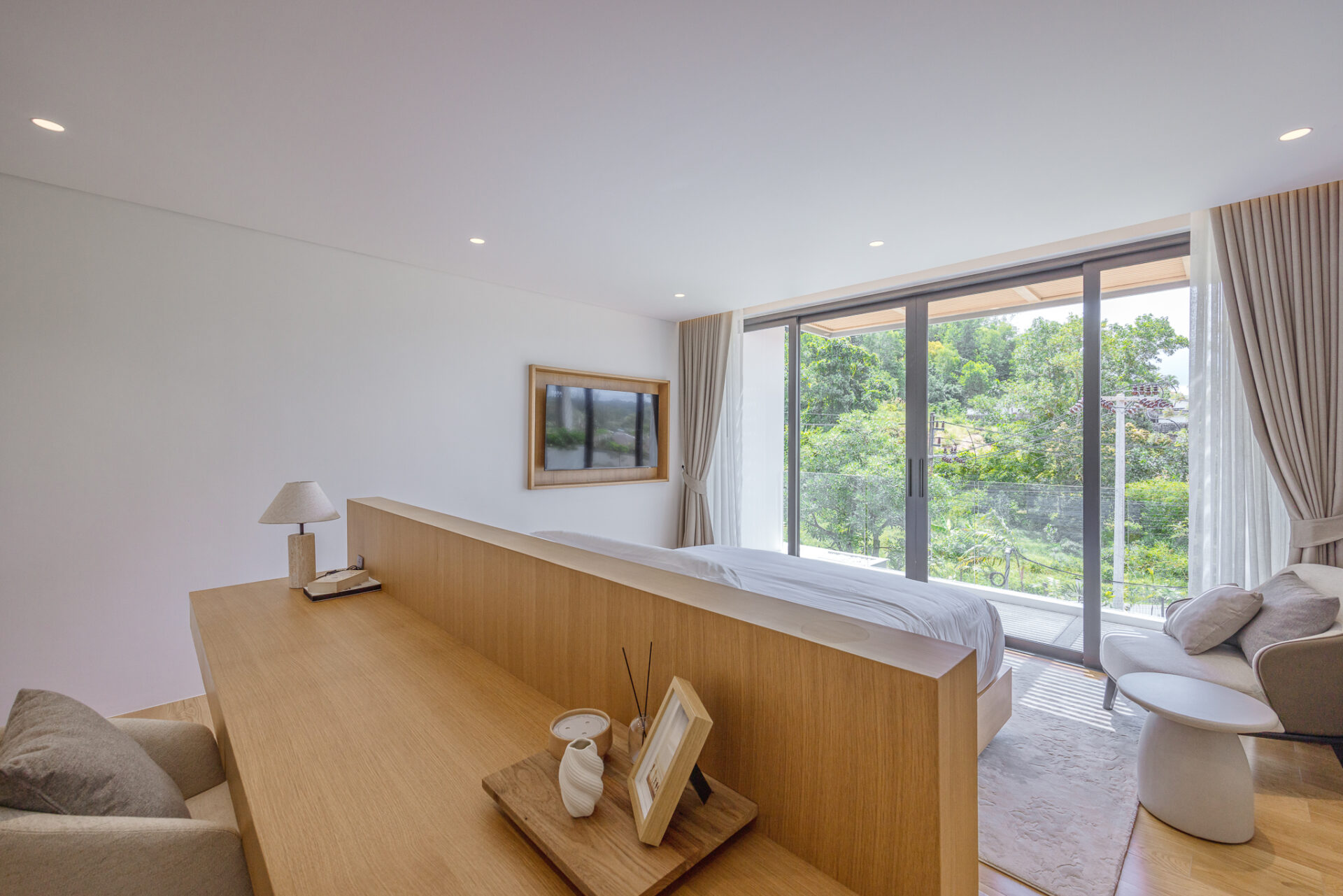 Modern bedroom with a wooden headboard/partition, bedside lamp, and large glass doors opening to a green garden outside.
