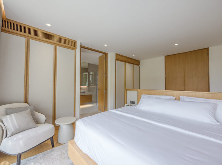 Hotel room with two white beds, light wood headboard, and built-in wardrobes; seating chair and small table by a window. Bedroom opens to a bathroom area visible through the doorway.