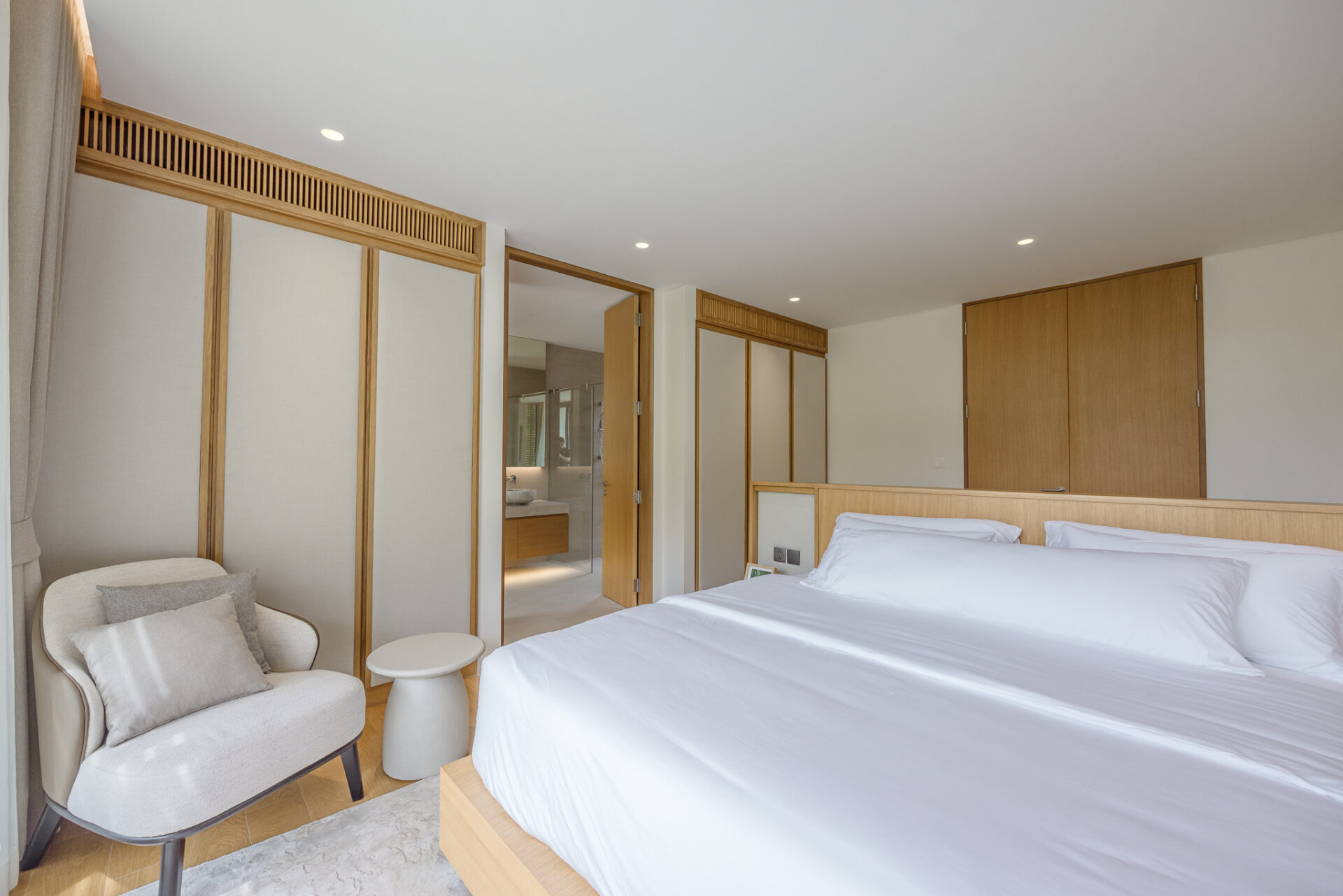 Hotel room with two white beds, light wood headboard, and built-in wardrobes; seating chair and small table by a window. Bedroom opens to a bathroom area visible through the doorway.