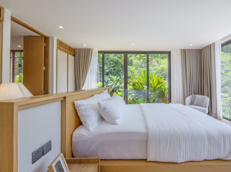 Bright hotel bedroom with a large white bed and a wooden headboard, open sliding doors to lush greenery outside.