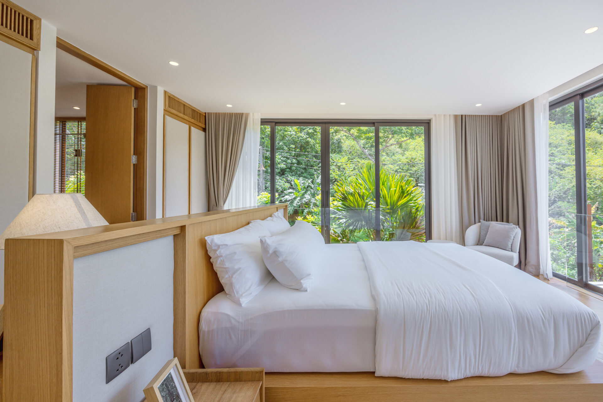Bright hotel bedroom with a large white bed and a wooden headboard, open sliding doors to lush greenery outside.