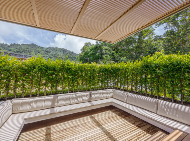 Outdoor seating area with a white cushioned L-shaped sofa on a wooden deck under a beige slatted pergola, overlooking green hedges and distant hills