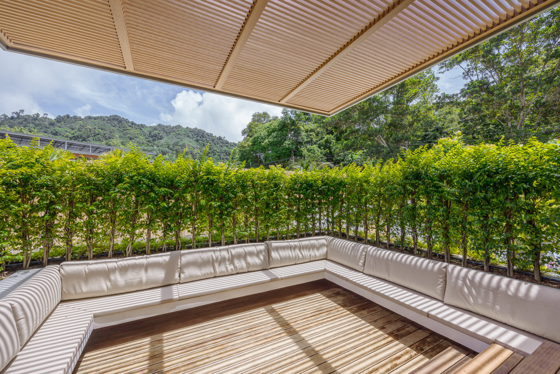 Outdoor seating area with a white cushioned L-shaped sofa on a wooden deck under a beige slatted pergola, overlooking green hedges and distant hills